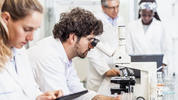 Ein Mann in einem Labor schaut durch ein Mikroskop, eine Frau schaut auf ein Tablet.